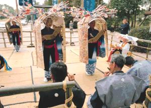 屋外の舞台上で、色鮮やかな飾りを冠した大きな笠を被り伝統衣装に身を包んで舞を披露する子供たちと、その手前で楽器を奏でながら儀式を支える正装した大人たちの姿を捉えた写真