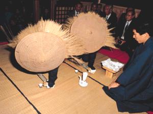 畳敷きの室内で大きな藁編みの笠を被った二人の人物が、長い竹の棒を床に打ち付け、その傍らで正装し静かに儀式を見守る男性たちの様子を捉えたカセドリ行事の写真