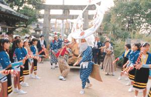 神社の石鳥居を背に、巨大な三日月形の被り物をした踊り手が中心で舞い、その両脇を鮮やかな青い法被を着て赤い飾りのついた棒を持つ子供たちが囲んで伝統的な舞を披露している浮立玄蕃一流の写真