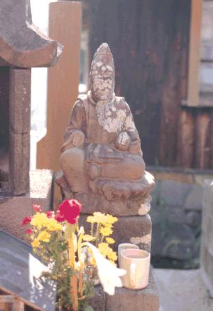 風化が進んだ石像が小祠の脇で穏やかに座し、手前には色鮮やかな供花と湯飲みが丁寧に供えられている様子の写真