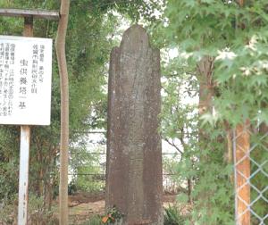 木々が生い茂り周囲を草木が覆う一角に風雨で古びた石碑が静かに佇み、その左側には由来を示す説明板が立つ様子の写真