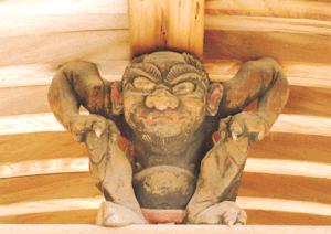 木造の梁下に据えられた鬼形の力強い彫刻が腕を張り膝を抱えて踏ん張る姿で緊張感ある造形美と迫力を放つ写真