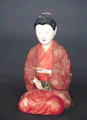 赤色を基調とした華やかな和装をまとい正座して器物を手にする人形が精緻な文様と布地の質感を示し落ち着いた姿で佇む写真
