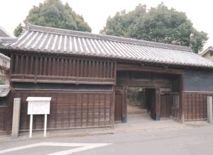道路に面して建つ、切妻造りの瓦屋根が載った重厚な木造の長屋門で、門扉の左側には格子窓が並び、右側には通用門と思われる小さな入り口が設けられた武家屋敷の門の外観写真