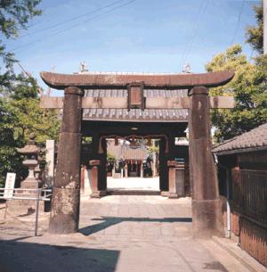 石畳の参道の入り口に建つ重厚な石造りの鳥居と、その奥に構える瓦屋根の大きな山門、さらにその先に見える社殿までが一直線に並んでいる石造肥前鳥居の外観写真