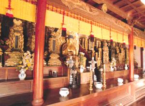 寺院の内陣で、中央に鎮座する黄金の仏像と左右に並ぶ数多くの位牌が、赤い柱や黄色い御簾に彩られた荘厳な空間の中で祀られ、手前の供物台には白い香炉や花が添えられた御位牌所の写真