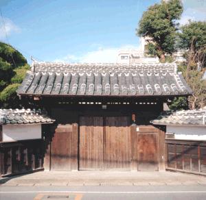 重厚な瓦屋根を載せた木造の大きな門が中央に構え、その両脇から白壁と黒い木枠の塀が左右に延びる、武家屋敷の門を正面から捉えた写真