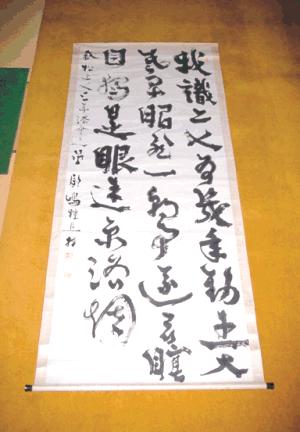 茶褐色の床に置かれた白い縦長の掛け軸に、勢いのある筆致の墨書きで漢字が三行にわたって力強く書かれており、左端には小さな文字の署名と朱色の印影が添えられている副島種臣の書の写真
