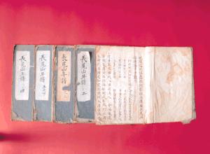 鮮やかな赤色の背景の上に、青色の表紙に白抜きの題箋が貼られた古い冊子が複数冊並べられ、そのうちの一冊が開かれて緻密な漢字が並ぶ本文が露出している長尾山年譜の写真