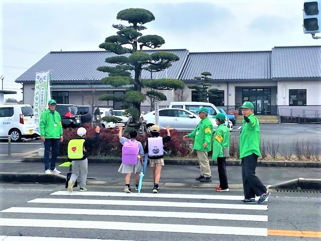 横断歩道を渡るランドセルを背負った3人の小学生に対し、緑色のジャンパーを着た見守り隊の大人たちが笑顔で手を振ったり周囲の安全を確認したりして、登下校の交通安全をサポートしている風景を捉えた写真