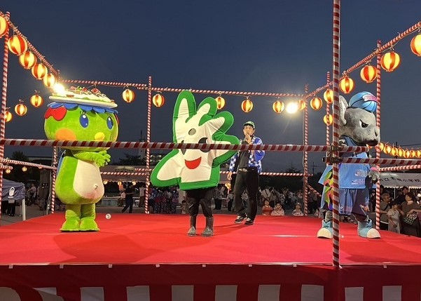 夕暮れの祭り会場で赤い櫓の舞台に三体の着ぐるみと司会者が並び、提灯の明かりに包まれながら盛り上げる姿が印象的に映し出されている写真