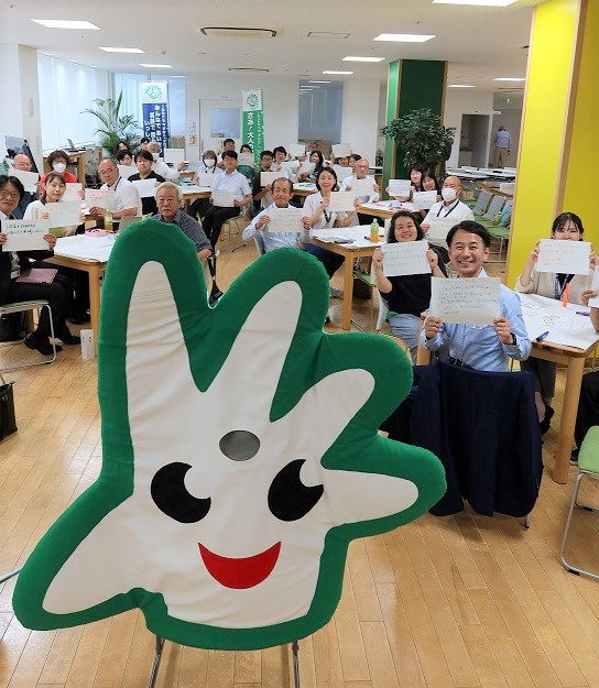 教室風の会場で多くの参加者が紙を掲げて座る中、前方に白緑の手型キャラクターが立つ様子を写した写真