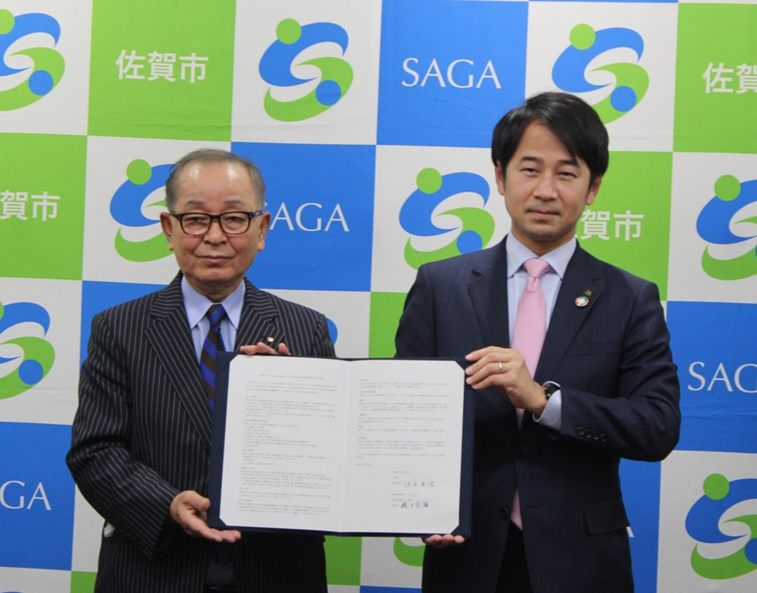 「SAGA」の文字と佐賀市章が格子状に並ぶバックパネルの前で、ストライプ柄のスーツを着た男性と無地のスーツにピンクのネクタイを締めた男性が、署名済みの協定書を共同で掲げ、組織間の強固な連携を象徴するように撮影された写真