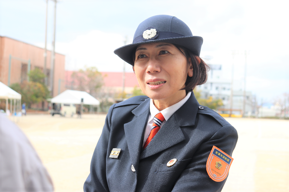 広場で、丸みのある紺色の帽子と左腕の袖には佐賀市消防団と記されたオレンジ色のワッペンが付いた制服を着用した園田由美さんがインタビュアーと話している様子の写真
