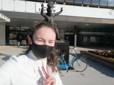 躍動感のある銅像を背景に、白いシャツを着て黒いマスクを着用した女性が、ピースサインをしながら晴れやかな笑顔の自撮り写真