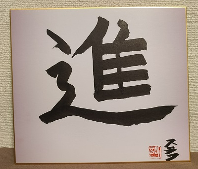 白い色紙の中央に、力強く伸びやかな筆致で「進」という一文字が墨で書かれ、右下には赤い落款と署名が添えられている写真