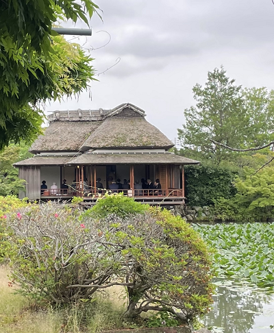 蓮の葉が浮かぶ池のほとりに建つ茅葺き屋根の歴史的な日本家屋に、多くの人物が座っているのを写した神野公園の隔林亭の外観写真