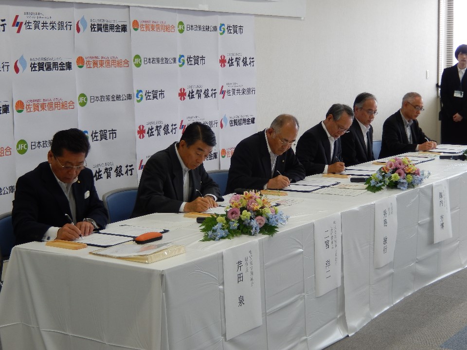 佐賀銀行や日本政策金融公庫、佐賀市などのロゴが並ぶパネルを背景に、白い布がかけられた長い机に座ったスーツ姿の男性6名が、一斉に手元の協定書へ署名を行っている様子の写真