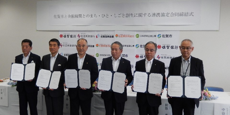 「佐賀市と金融機関とのまち・ひと・しごと創生に関する連携協定合同締結式」の横断幕を背景に、署名済みの協定書を開いて掲げたスーツ姿の男性代表者六名が、横一列に並んで記念撮影に応じている写真