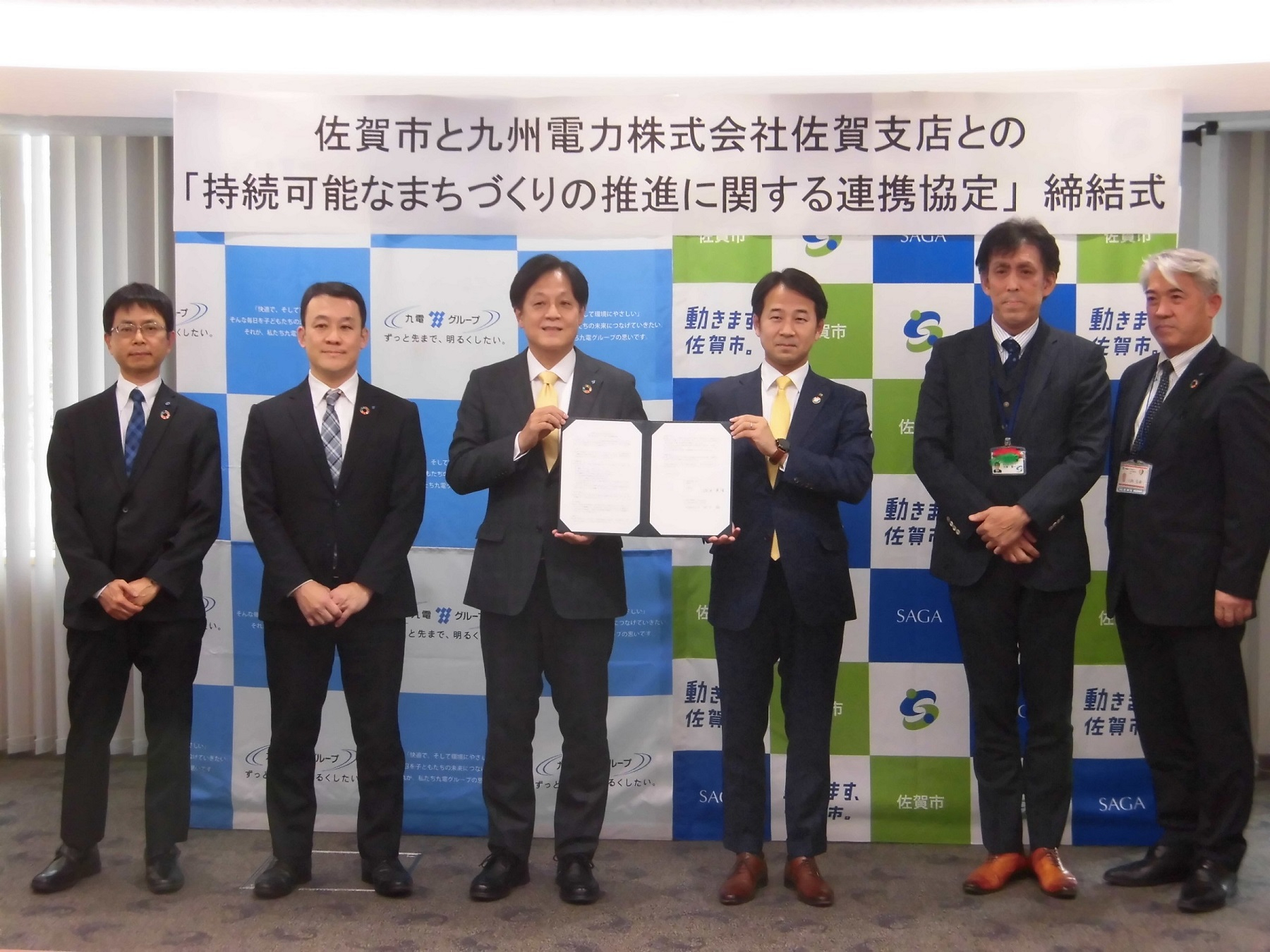 佐賀市のロゴ入りバックパネル前で、スーツ姿の男性6名が並び、中央の2名が協定書を掲げている、佐賀市と九州電力株式会社佐賀支店との「持続可能なまちづくりの推進に関する連携協定」締結式の様子の写真