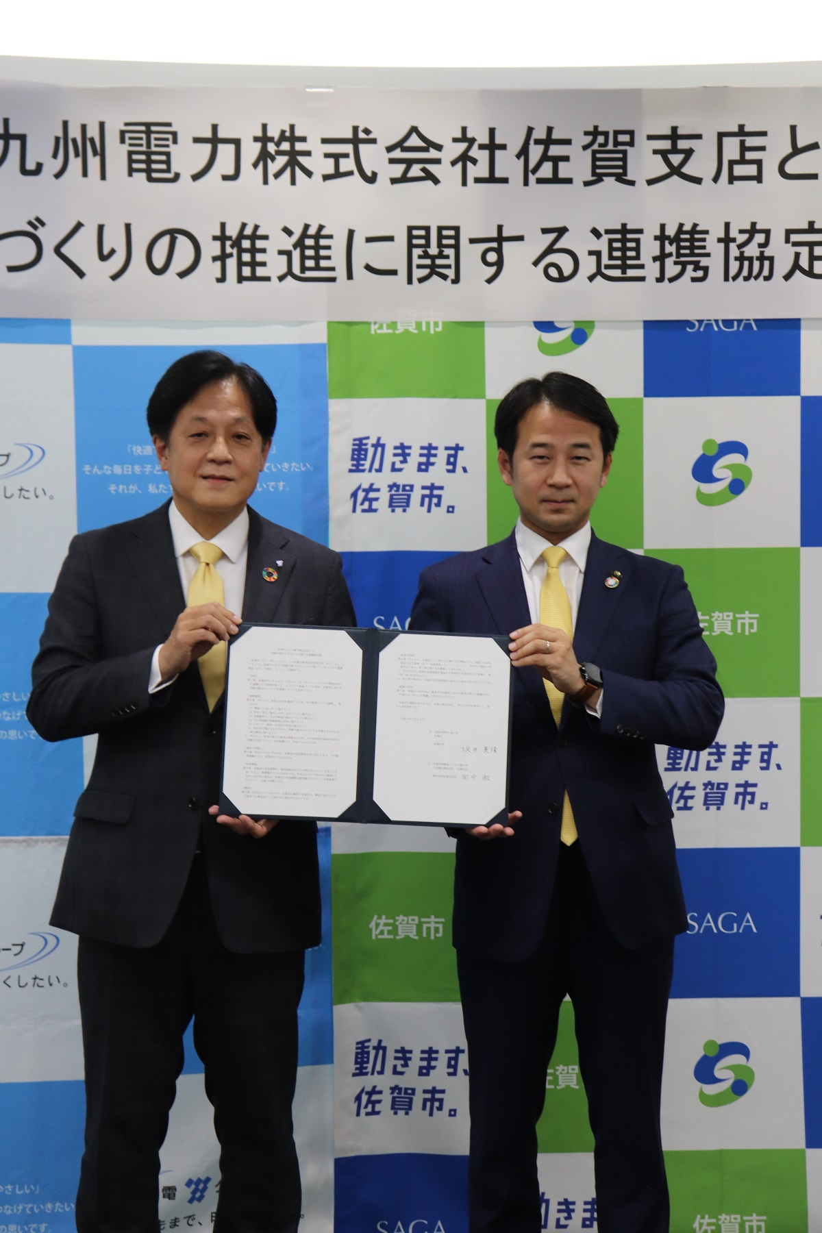 佐賀市のロゴや「動きます、佐賀市。」という文字が並ぶバックパネルが設置されている背景の前に、スーツ姿の二人の男性が協定書を広げて持ち並んでいる、九州電力株式会社佐賀支店と佐賀市の連携協定締結式の様子の写真