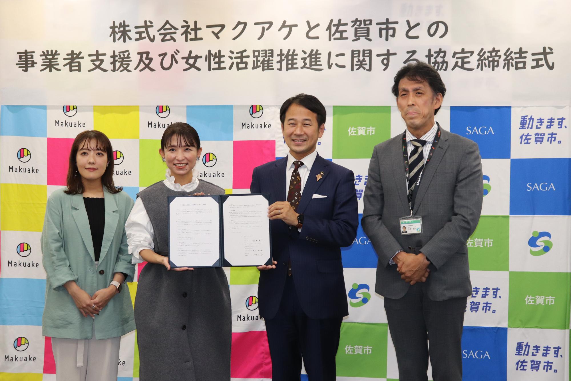 株式会社マクアケと佐賀市のロゴが並ぶバックボードの前で、協定書を持つ中央の男女二人に加え、左側にミントグリーンのジャケットを着た女性、右側にグレーのスーツを着た男性が並んで笑顔を見せる締結式での集合写真