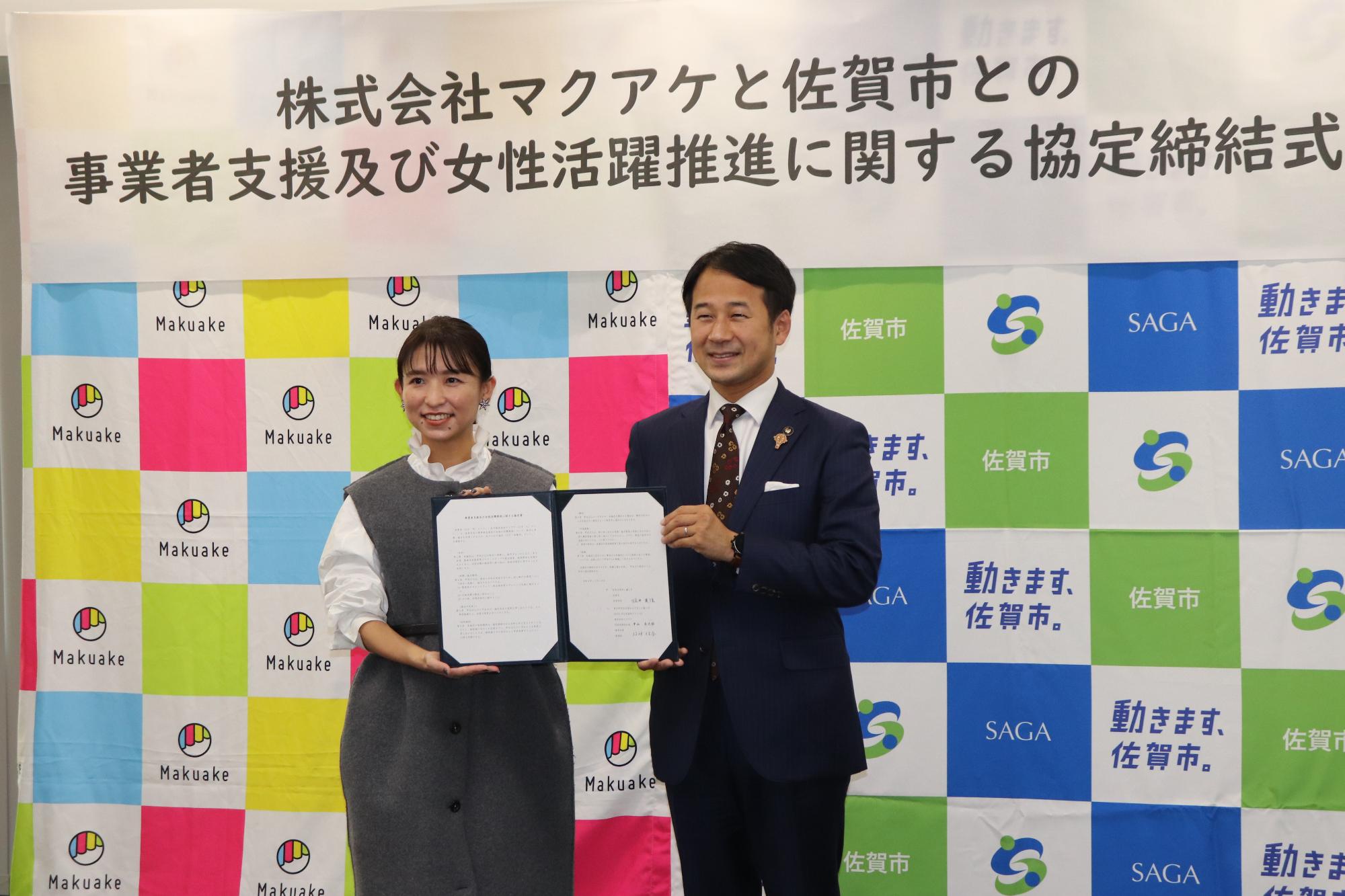 株式会社マクアケと佐賀市のロゴが敷き詰められたバックボードを背に、グレーのワンピースを着た女性と紺色のスーツを着た市長が、見開き状態の協定書を二人で持ちながら笑顔で並んでいる、協定締結式での記念写真