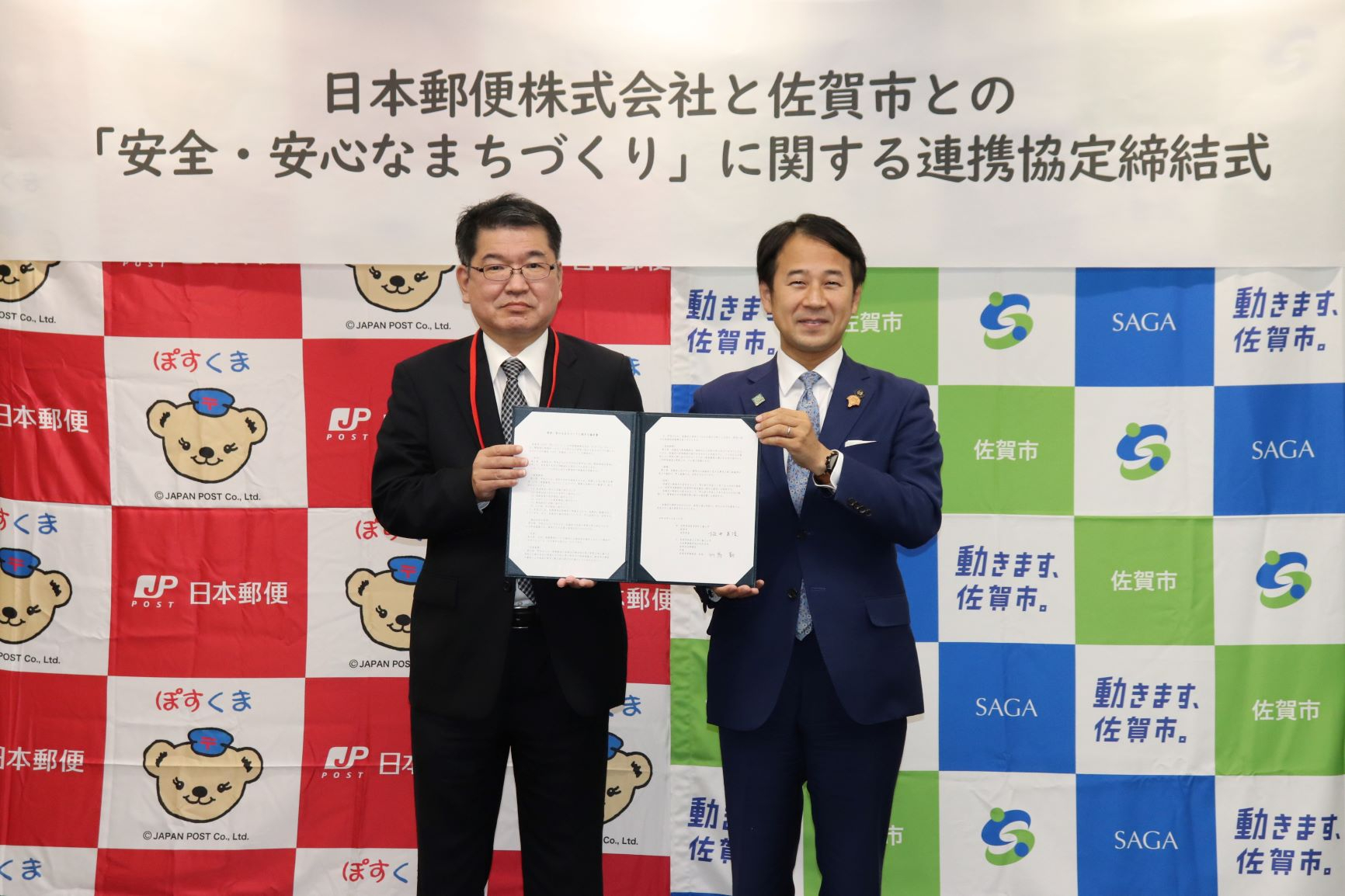 日本郵便のキャラクターや佐賀市のロゴが並ぶバックパネル前で、スーツ姿の男性2名が並び、開いた協定書を中央で一緒に掲げている、日本郵便株式会社と佐賀市との「安全・安心なまちづくり」に関する連携協定締結式の記念写真