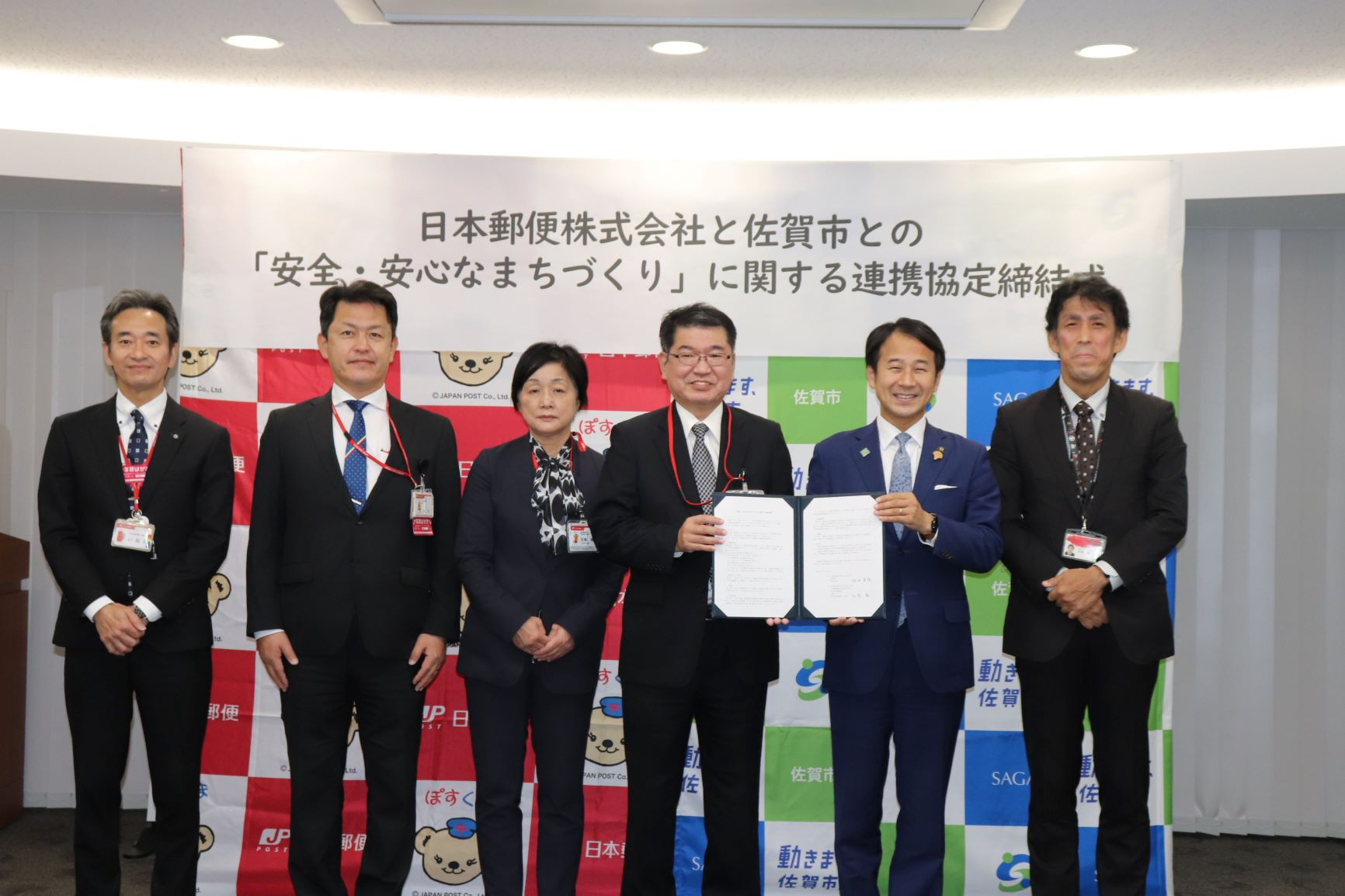 日本郵便のキャラクターや佐賀市のロゴが並ぶバックパネル前で、スーツ姿の男女6名が並び、中央の2名が開いた協定書を掲げている、日本郵便株式会社と佐賀市との「安全・安心なまちづくり」に関する連携協定締結式の様子の写真