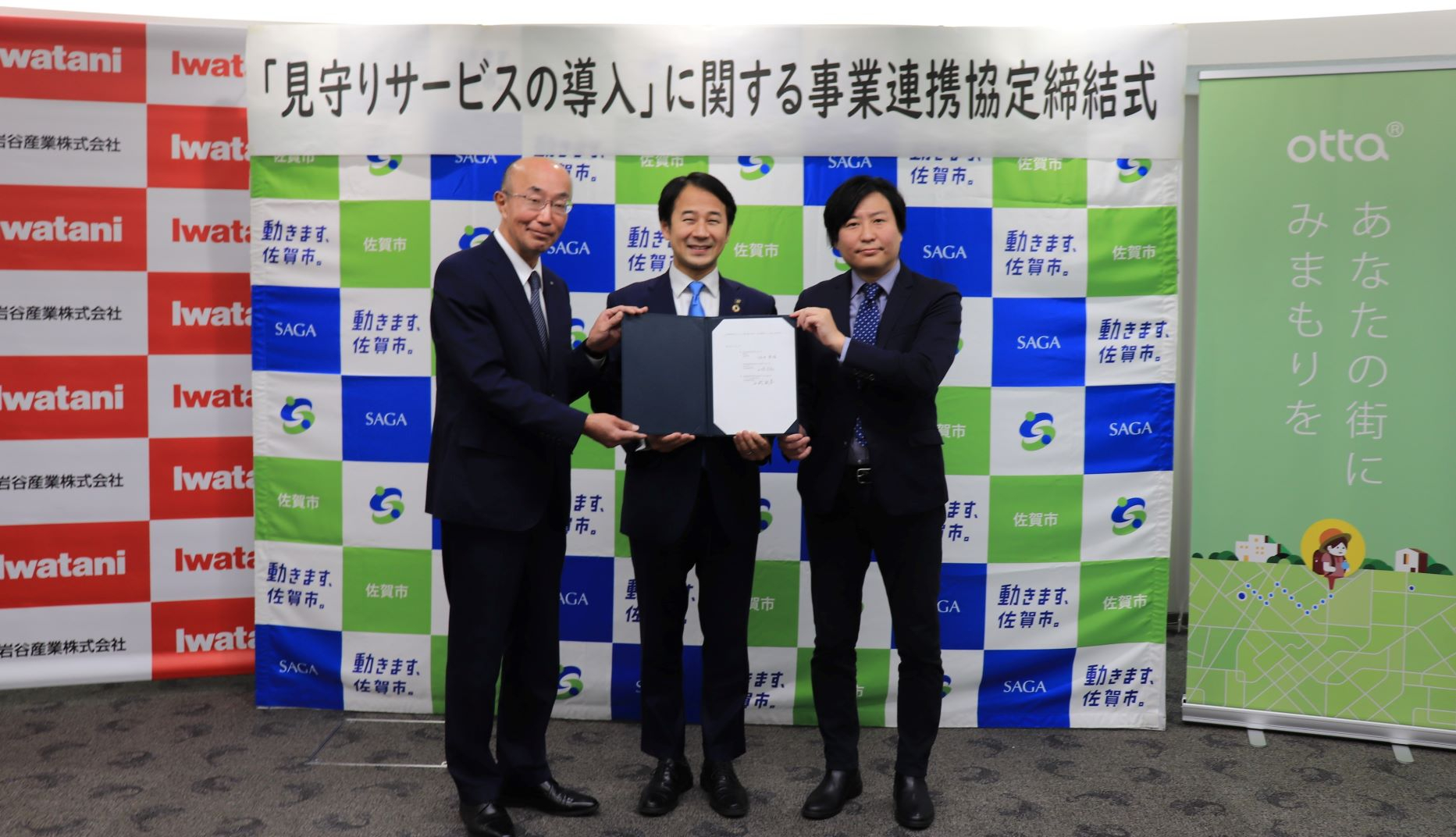 「見守りサービスの導入」に関する事業連携協定締結式の横断幕を背景に、3者のロゴが並ぶボードの前で、スーツ姿の男性三人が開いた状態の協定書を共同で持ちながら笑顔を見せている記念写真