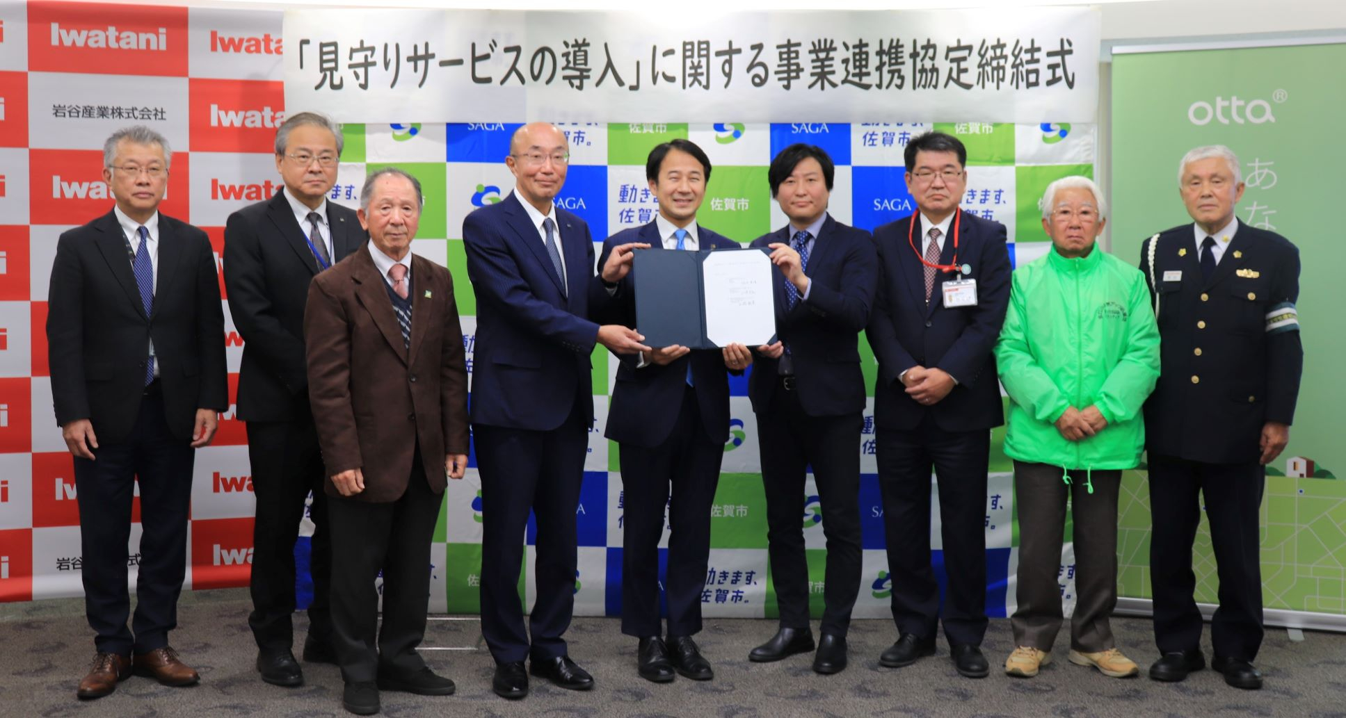 「見守りサービスの導入」に関する事業連携協定締結式の横断幕を背景に、3者のロゴが並ぶボードの前で、協定書を持つ中央の男性三人を囲むように、警察官や緑のジャンパーを着た男性ら関係者九名が並んだ記念写真