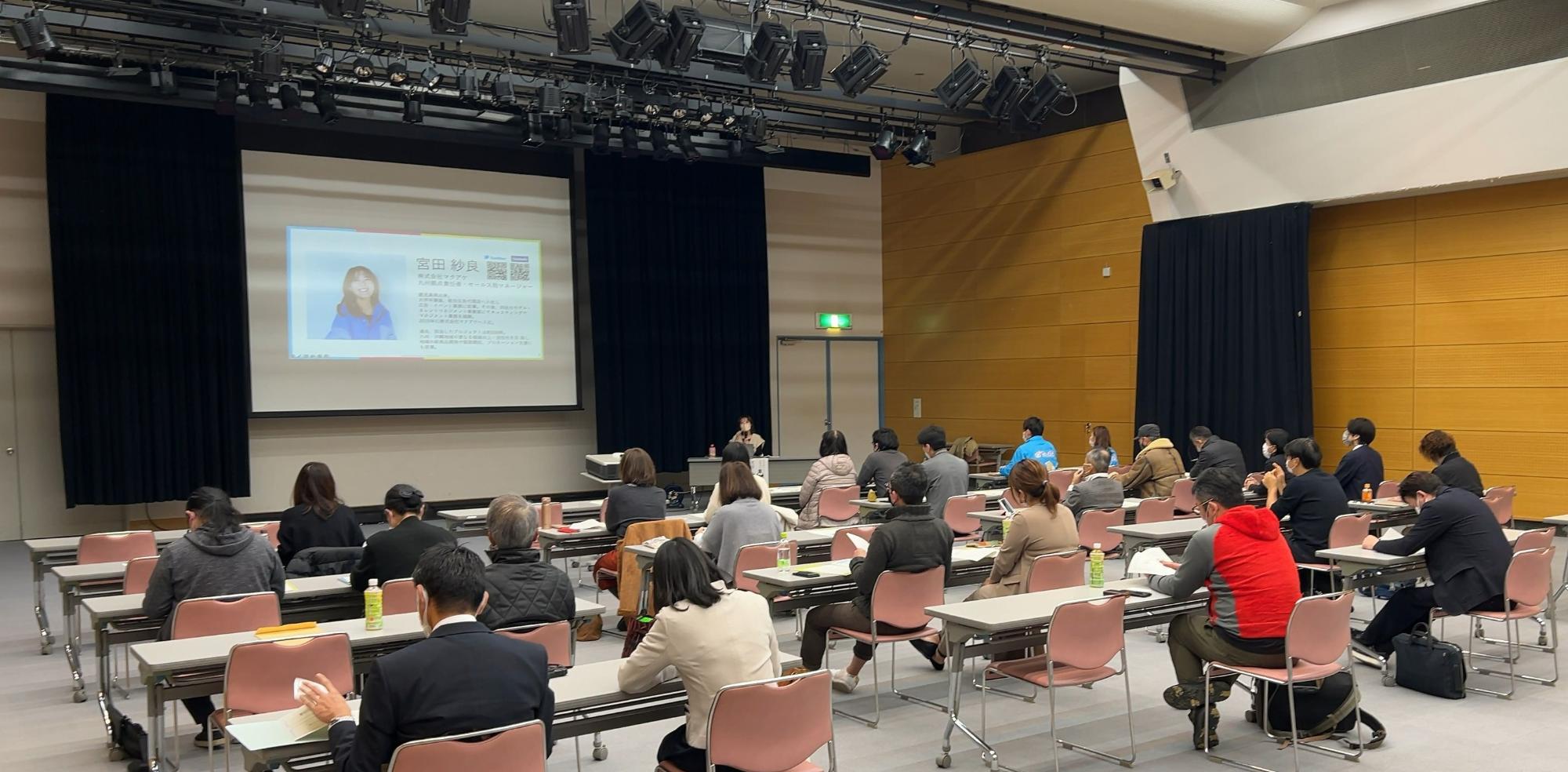 前方に講師の紹介スライドが投影された大きなスクリーンがあるホールにて、間隔を空けて配置された机に座った多くの受講生たちが、前方で行われている講義を静かに聴講している様子を後方から捉えた写真