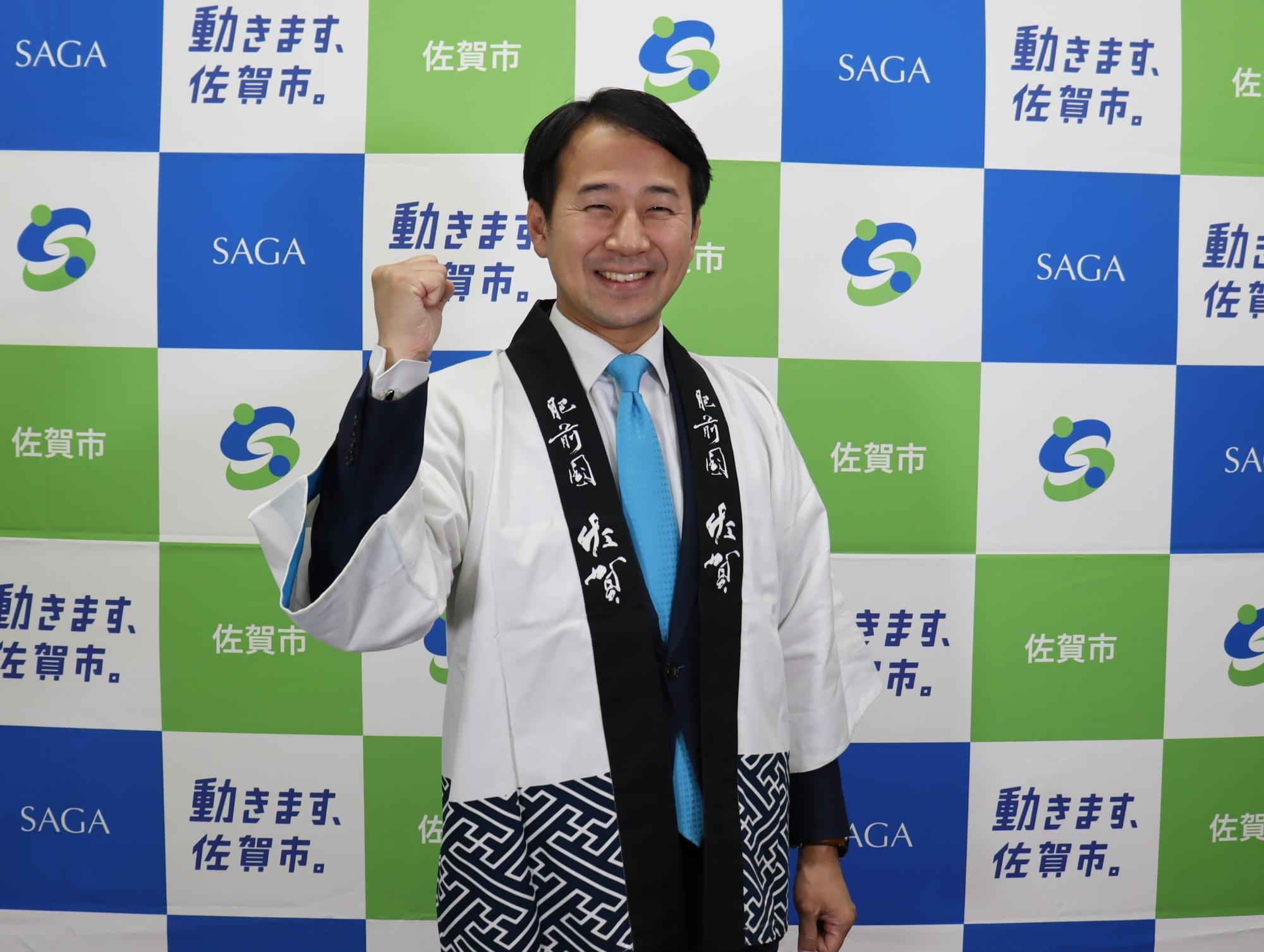 「動きます、佐賀市。」というキャッチコピーや市章が並ぶ青と緑の格子模様の背景パネルの前で、白地に黒い襟の法被を羽織りネクタイを締めた市長が、晴れやかな笑顔で右手の拳を力強く握りしめるガッツポーズをした写真