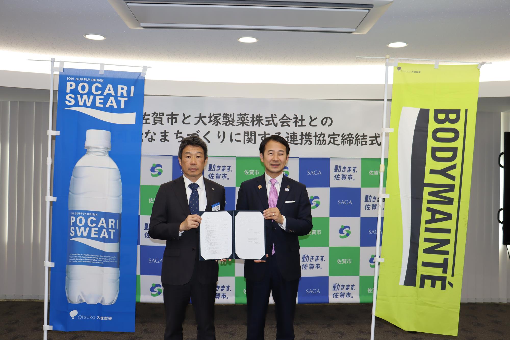 左右にはポカリスエットとボディメンテの大きなのぼり旗が設置されており、佐賀市のロゴパネルが設置された背景の前で、佐賀市と大塚製薬株式会社との連携協定締結式にて、スーツ姿の男性2名が協定書を手に並んでいる記念写真