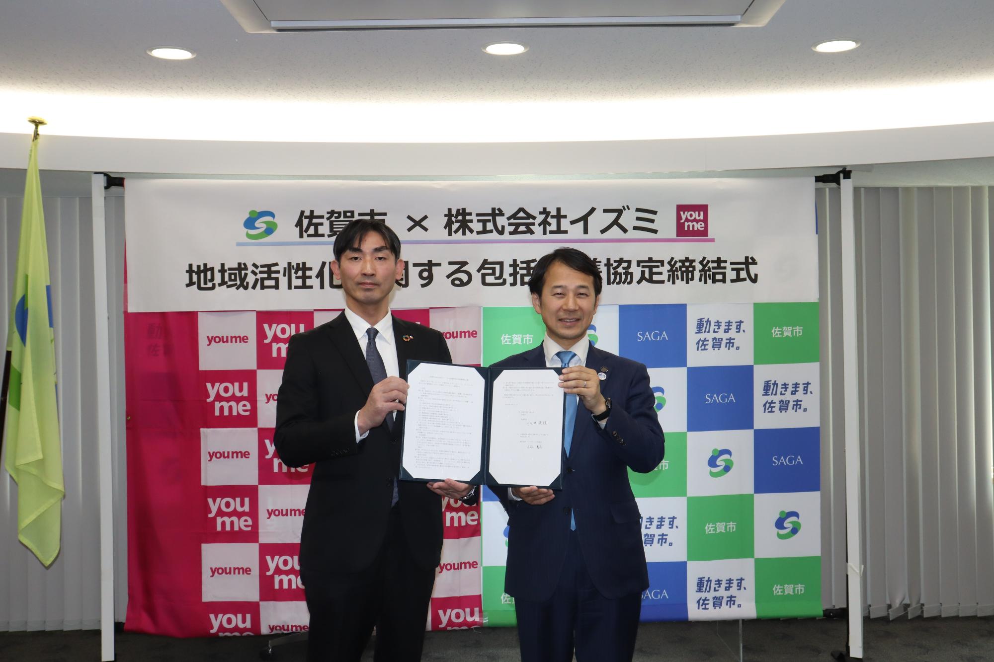 佐賀市と株式会社イズミのロゴが描かれた横断幕を背景に、スーツ姿の男性二人が地域活性化に関する包括連携協定締結式の協定書を手に持ち、正面を向いて並んでいる締結式の様子を捉えた記念写真
