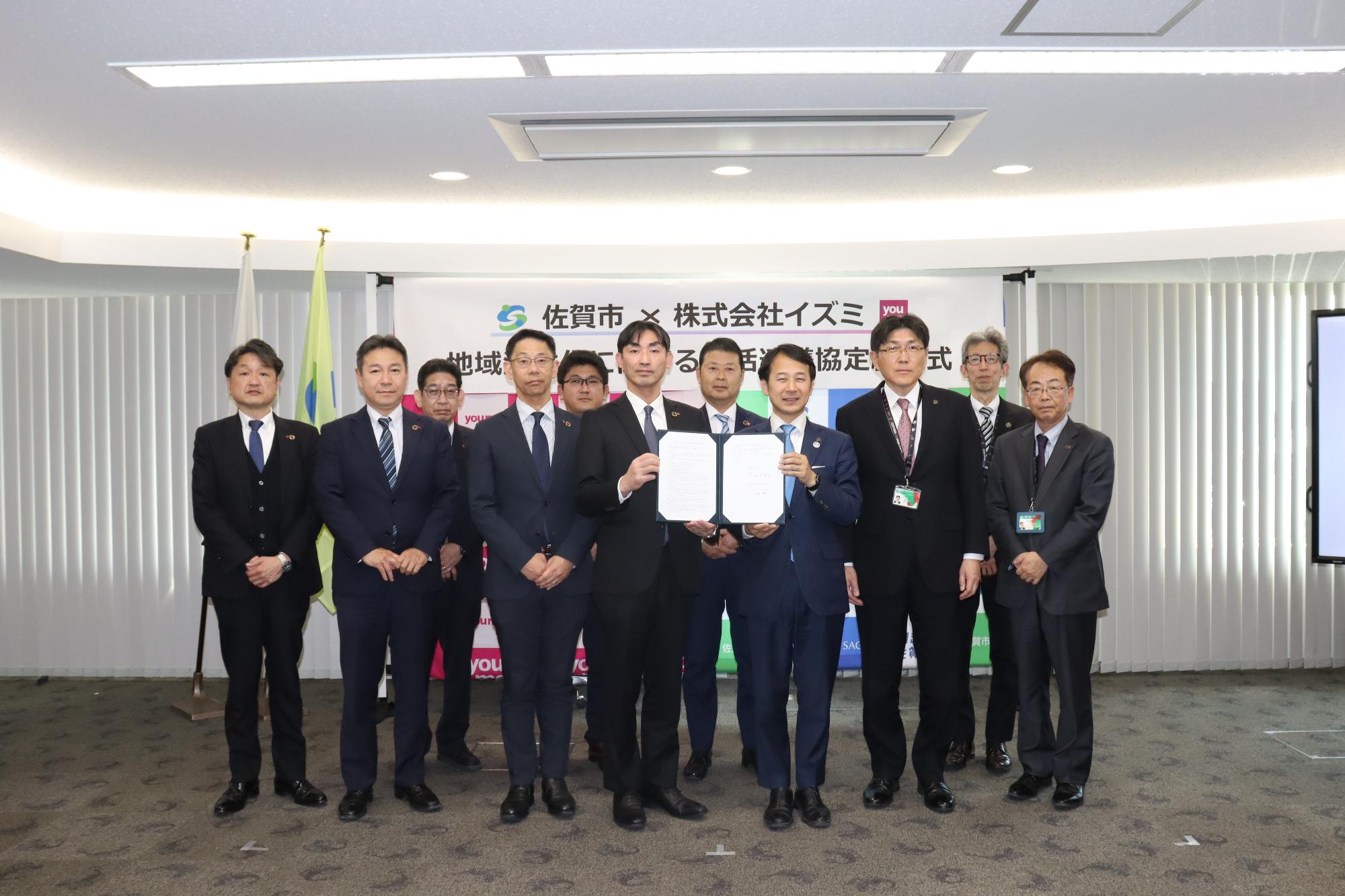 佐賀市と株式会社イズミのロゴが記された地域活性化包括連携協定締結式の横断幕を背に、協定書を掲げた中央の男性二人を含むスーツ姿の関係者11名が、室内で一列に並んでカメラを見つめている集合写真
