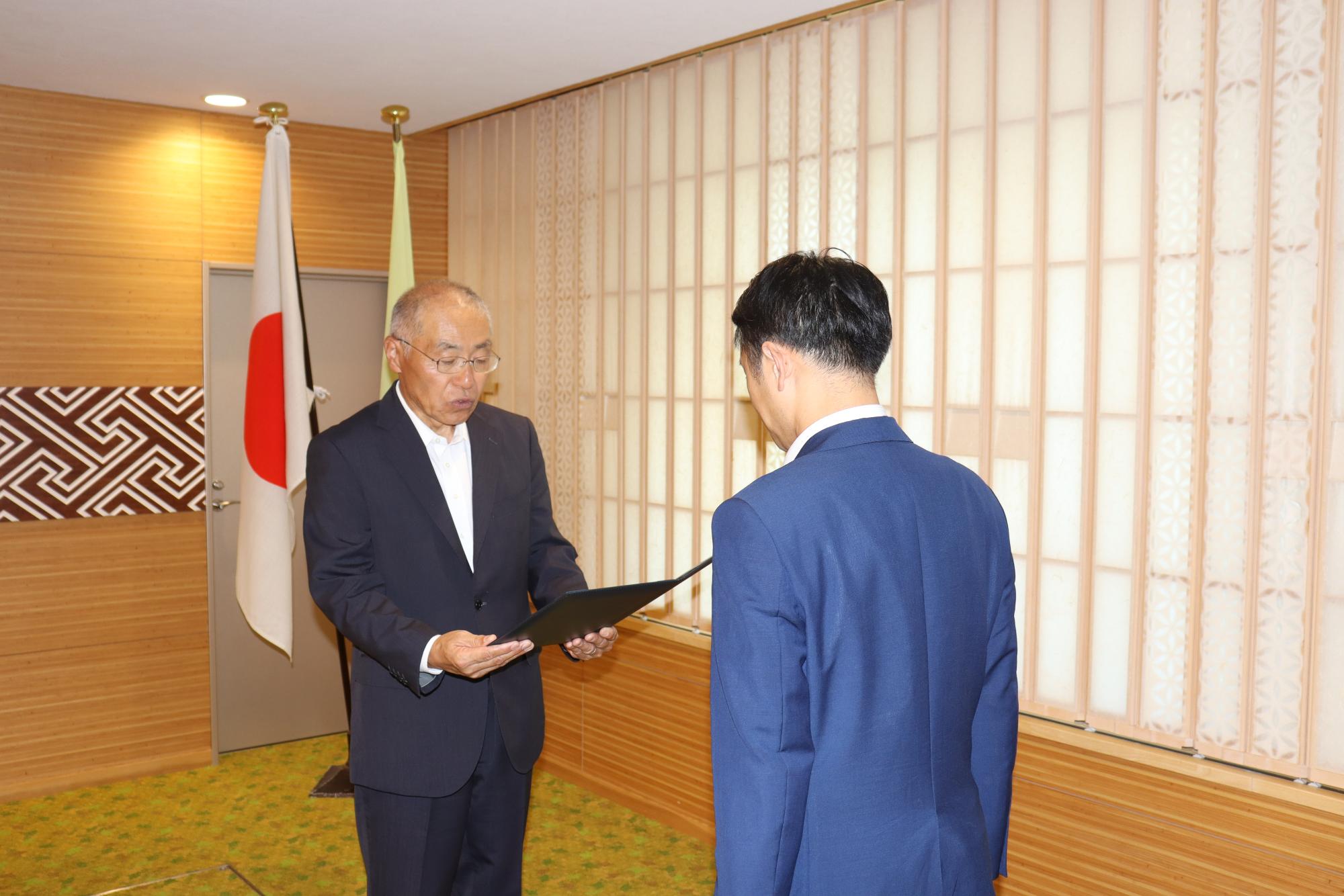 木目調の壁と日本の国旗がある室内で、眼鏡をかけたスーツ姿の男性が書類を手に持ち、背を向けた青いスーツの男性に対し答申が行われている様子の写真