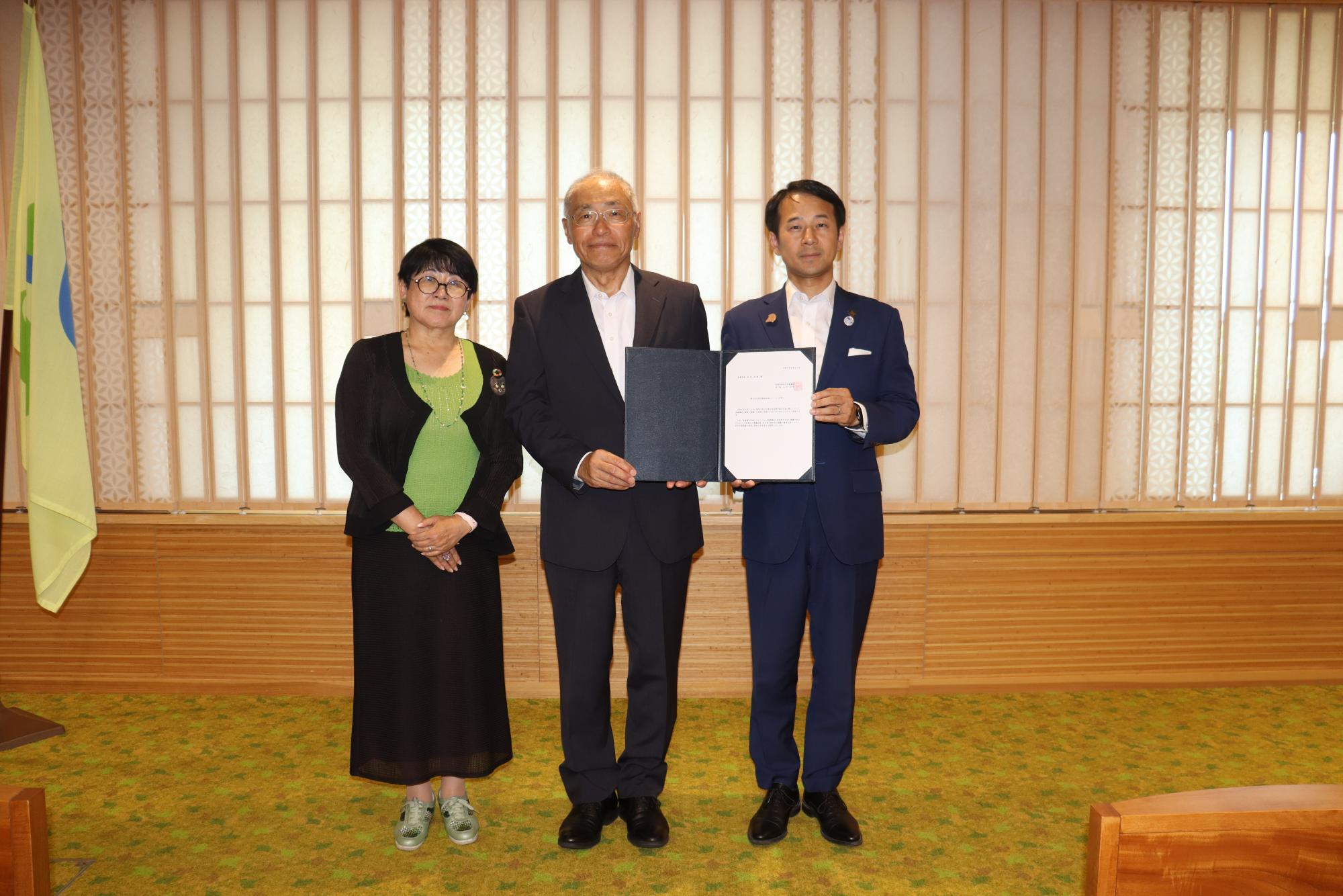 純和風の室内で、スーツ姿の男性二人が見開いた証書を中央で一緒に持ち、その左隣に緑色のインナーと黒いスーツ姿の女性が並んで立っている写真