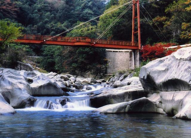 緑豊かな山間を流れる清流に大きな岩が並び、その上を赤い吊り橋が架かっている風景の中で、手前の川面と奥の木々や一部紅葉した葉が美しいコントラストを描いている、自然豊かな渓谷を捉えた雄淵雌淵の写真
