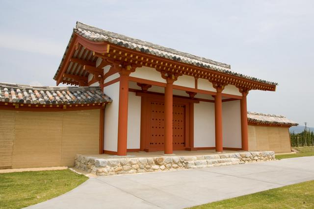 薄曇りの空の下で、朱色の柱と白い壁が特徴的な伝統的な日本建築の門が石積みの土台の上に建っており、瓦屋根が左右の土壁へと続く、平坦な道と芝生に囲まれた静かな肥前国庁跡南門の外観を捉えた写真