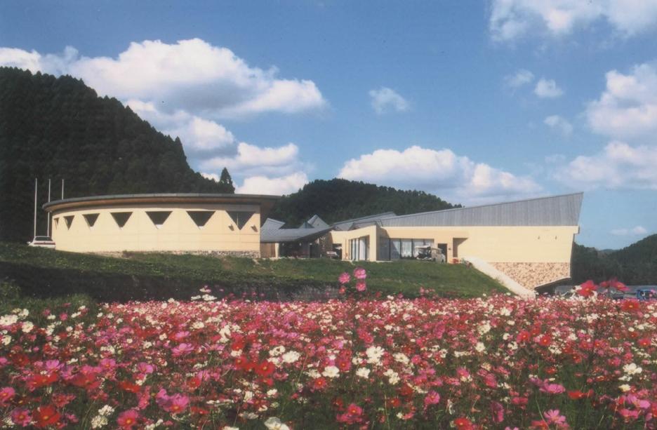 青空の下、手前に広がる色鮮やかなピンクや白のコスモス畑の向こう側に、円形や鋭角な屋根を持つモダンなデザインのベージュ色のやまびこの湯の建物が建ち、背景には深い緑の山々が連なる、自然と建築が調和した風景を捉えた写真