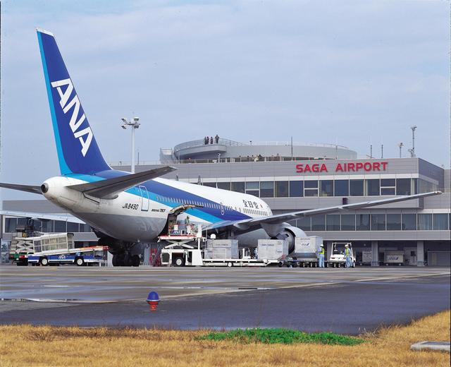空港の駐機場で、垂直尾翼に「ANA」のロゴが描かれた青と白の旅客機が停車しており、背景には「SAGA AIRPORT」と書かれたターミナルビルや作業車両、手配を行うスタッフの姿が見える、活気ある九州佐賀国際空港の風景を捉えた写真