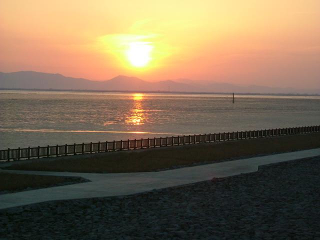 オレンジ色に染まる夕空の下、穏やかな海面に夕日の光が反射して一筋の輝きを作り出し、遠くには幾重にも重なる山々のシルエットが浮かび、手前の遊歩道沿いの柵が静かな海岸線の風景を際立たせている、叙情的な夕暮れ時の干潟よか公園の写真