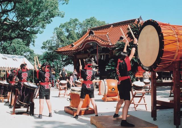 晴天の香椎神社境内で、お揃いの黒い法被を纏った若者たちが、大小様々な和太鼓を力強く打ち鳴らす伝統芸能のパフォーマンスを披露しており、背景の社殿や緑の木々が祭りの活気ある雰囲気を引き立てている、躍動感あふれる行事の風景を捉えた写真