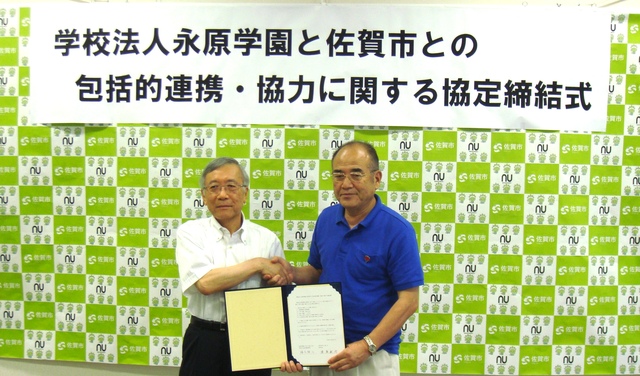 学校法人永原学園と佐賀市との包括的連携・協力に関する協定締結式の横断幕とバックパネルを背景に、白いシャツの男性と青いポロシャツの男性が、開いた協定書を手に持ちながら笑顔で握手を交わしている記念写真