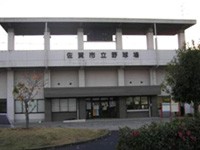 灰色の外壁を持つ平屋建ての公共施設で、正面玄関上部に佐賀市立野球場の看板が掲げられ、前面に低木の植栽がある建物外観の写真