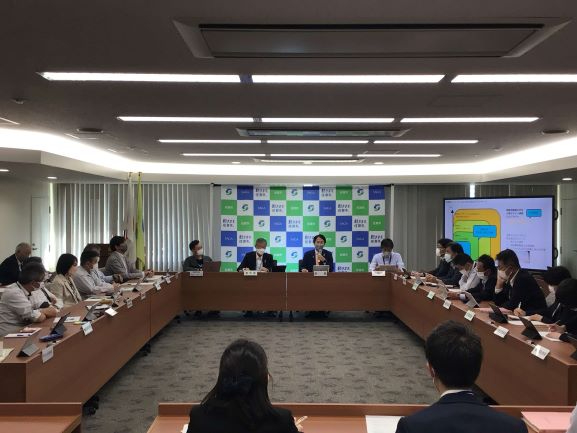 コの字型のテーブルを囲んで多くの出席者が会議を行っている会議室の様子で、正面には市松模様のバックパネルが置かれ、右奥にはスライドが表示されたモニターがあり、真剣な表情で話し合う人々を捉えた写真
