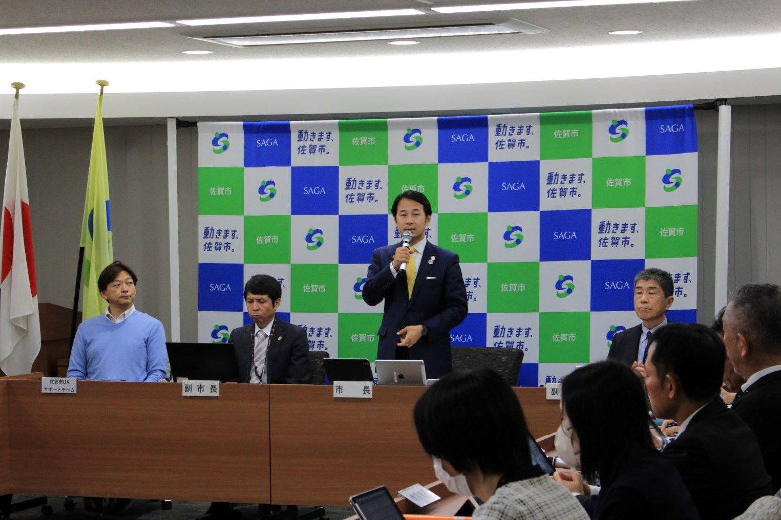 会議室で市長が立って説明を行い参加者が長机で資料を確認しながら会議を進める様子を壁面のパネル前で写した写真