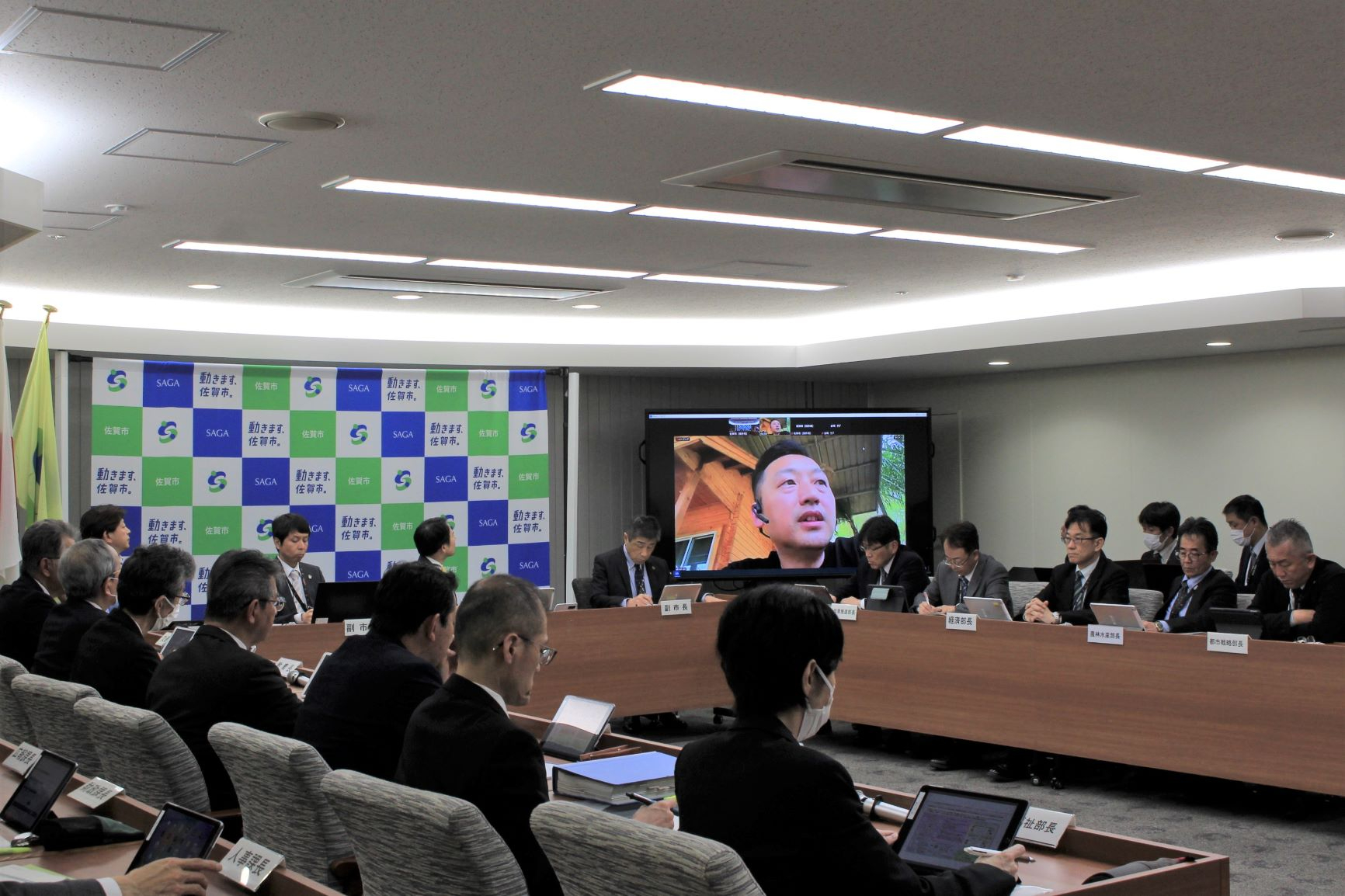 会議室で参加者が長机を囲み背景のパネル前で大型画面の映像を見ながら活発に議論を進める様子を捉えた写真