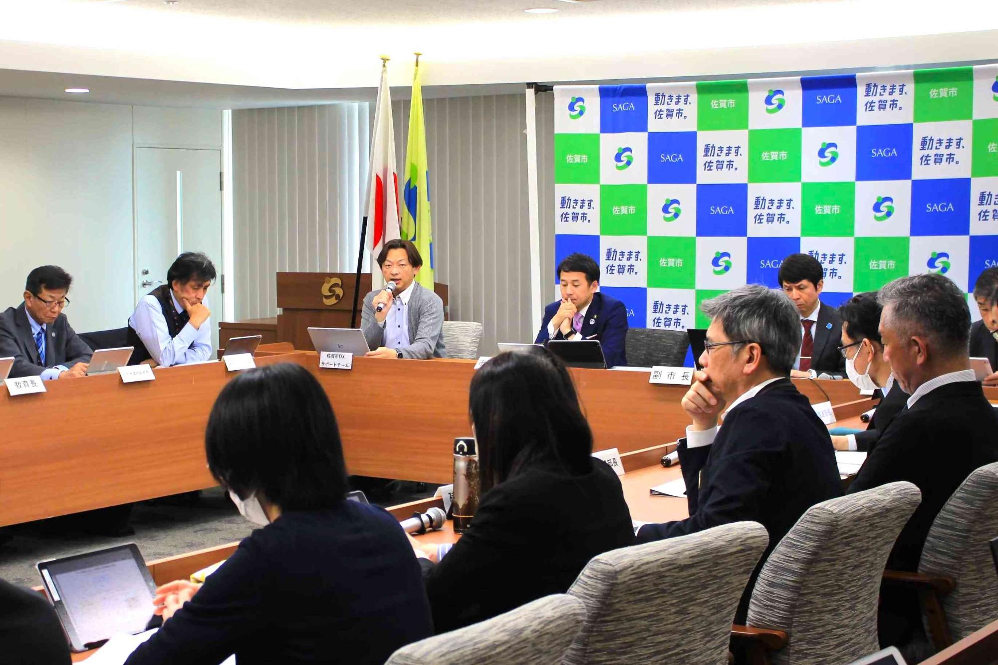 背景に旗とパネルが並ぶ会議室で参加者が長机を囲み資料や端末を確認しつつ議論を進める様子を写した写真
